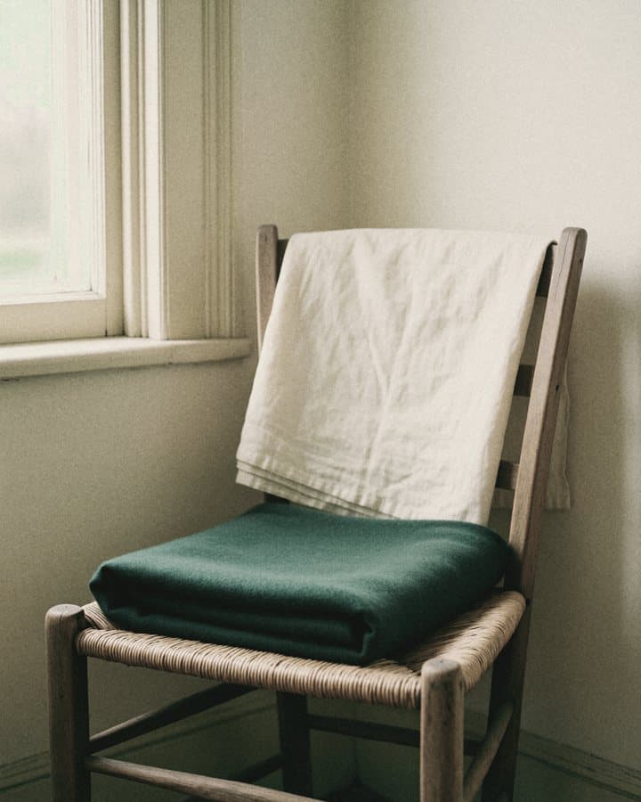 Folded ivory linen and a green wool blanket on a rush-seat chair by a window